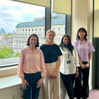 Four people stand by a window with a cityscape outside, smiling, wearing light-coloured smart-casual outfits.
