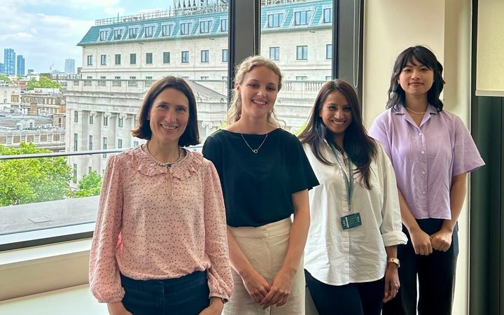 Four people stand by a window with a cityscape outside, smiling, wearing light-coloured smart-casual outfits.