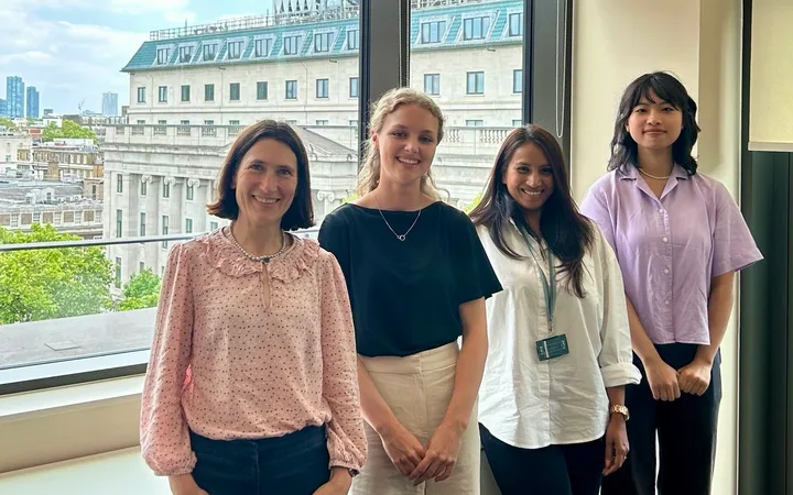 Four people stand by a window with a cityscape outside, smiling, wearing light-coloured smart-casual outfits.