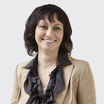 A smiling person with dark hair wearing a beige blazer and a black ruffled blouse against a plain grey background.