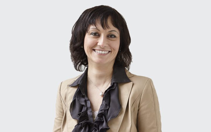 A smiling person with dark hair wearing a beige blazer and a black ruffled blouse against a plain grey background.