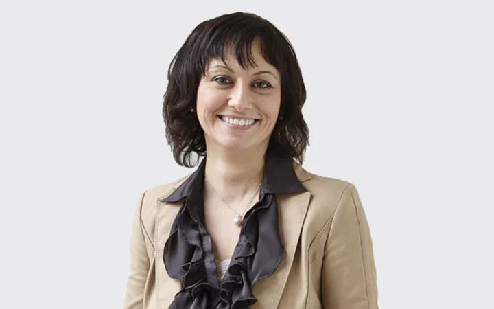 A smiling person with dark hair wearing a beige blazer and a black ruffled blouse against a plain grey background.