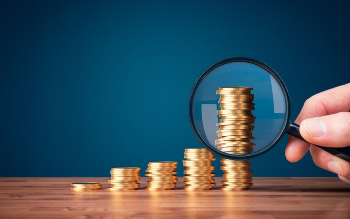 A hand holds a magnifying glass over stacks of gold coins arranged in increasing height on a wooden surface, set against a dark blue background.