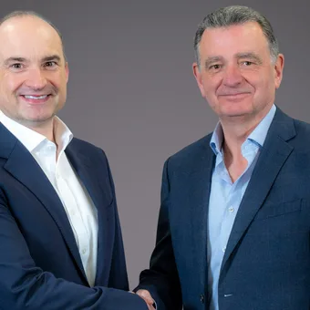 Two people in business suits shaking hands and smiling against a grey background.