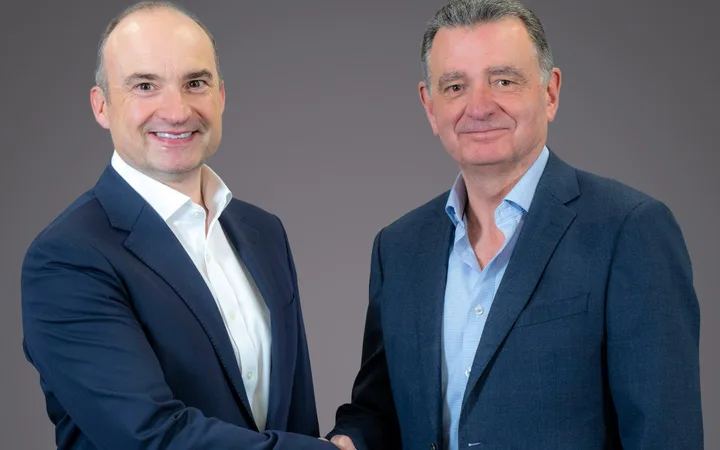Two people in business suits shaking hands and smiling against a grey background.