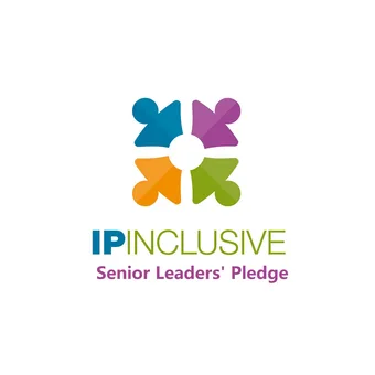 Portrait of a person with short grey hair, wearing a dark suit, light shirt and purple tie, facing the camera against a grey backdrop.