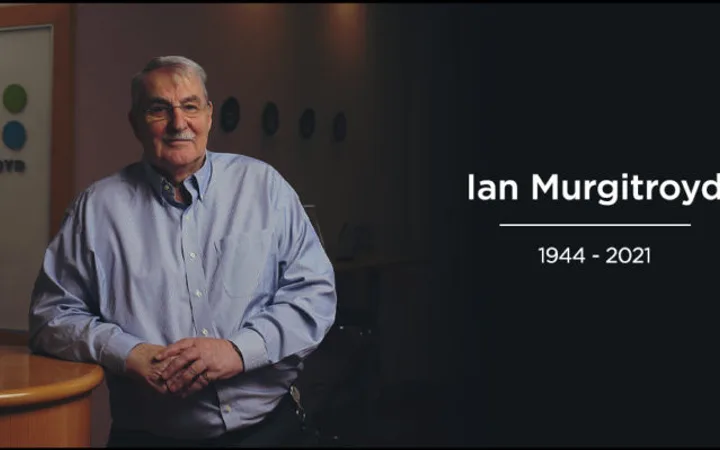 An elderly person in a blue shirt stands with hands clasped in front of a Murgitroyd sign, smiling in a professional setting, with a dark background.