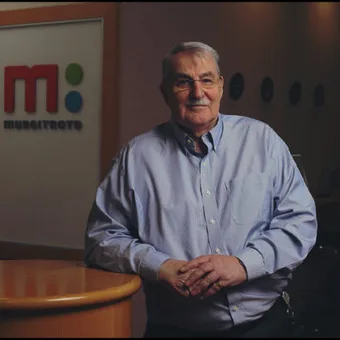 A mature person with glasses and a light blue shirt standing in an office with a Murgitroyd logo in the background.