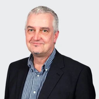 A middle-aged person with short grey hair, light beard, wearing a black blazer and checkered shirt, smiling against a light grey background.