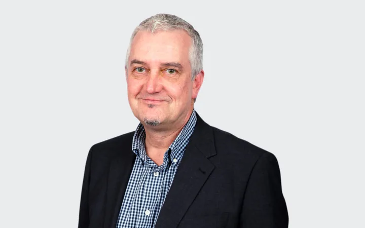 A middle-aged person with short grey hair, light beard, wearing a black blazer and checkered shirt, smiling against a light grey background.
