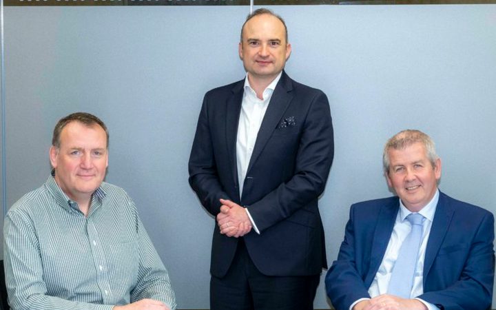 Three people in formal attire pose for a photograph in an office setting with a grey wall and a "m: m:" logo behind them.
