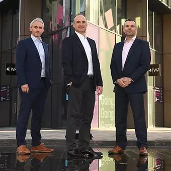Three people in suits standing in front of a modern office building with glass windows and digital displays.