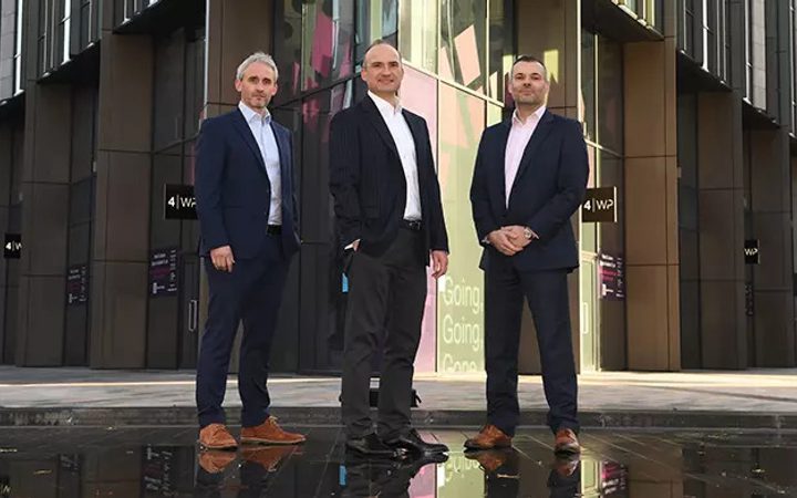 Three people in suits standing in front of a modern office building with glass windows and digital displays.
