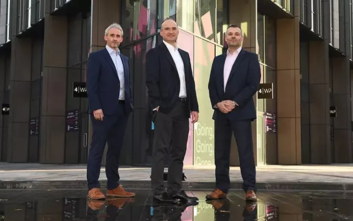 Three people in suits standing in front of a modern office building with glass windows and digital displays.