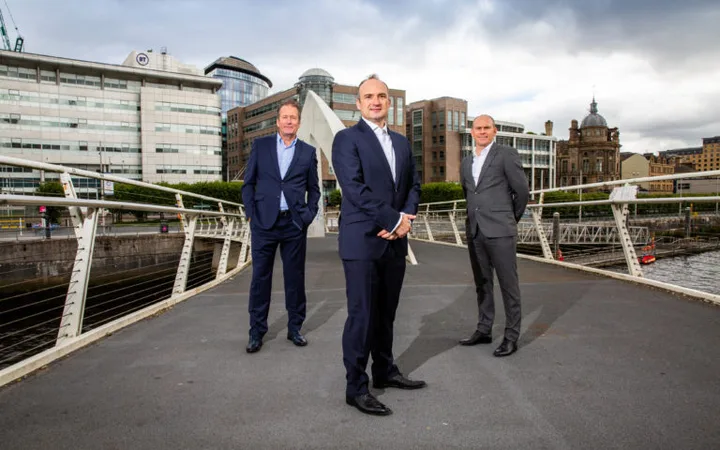 Three people in suits standing on a bridge in an urban area with modern buildings and cloudy sky in the background.