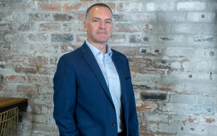 Person in a blue suit standing against an exposed brick wall with a neutral expression.