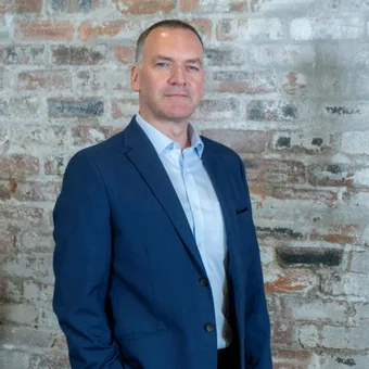 A person in a blue suit standing against a brick wall with a serious expression.
