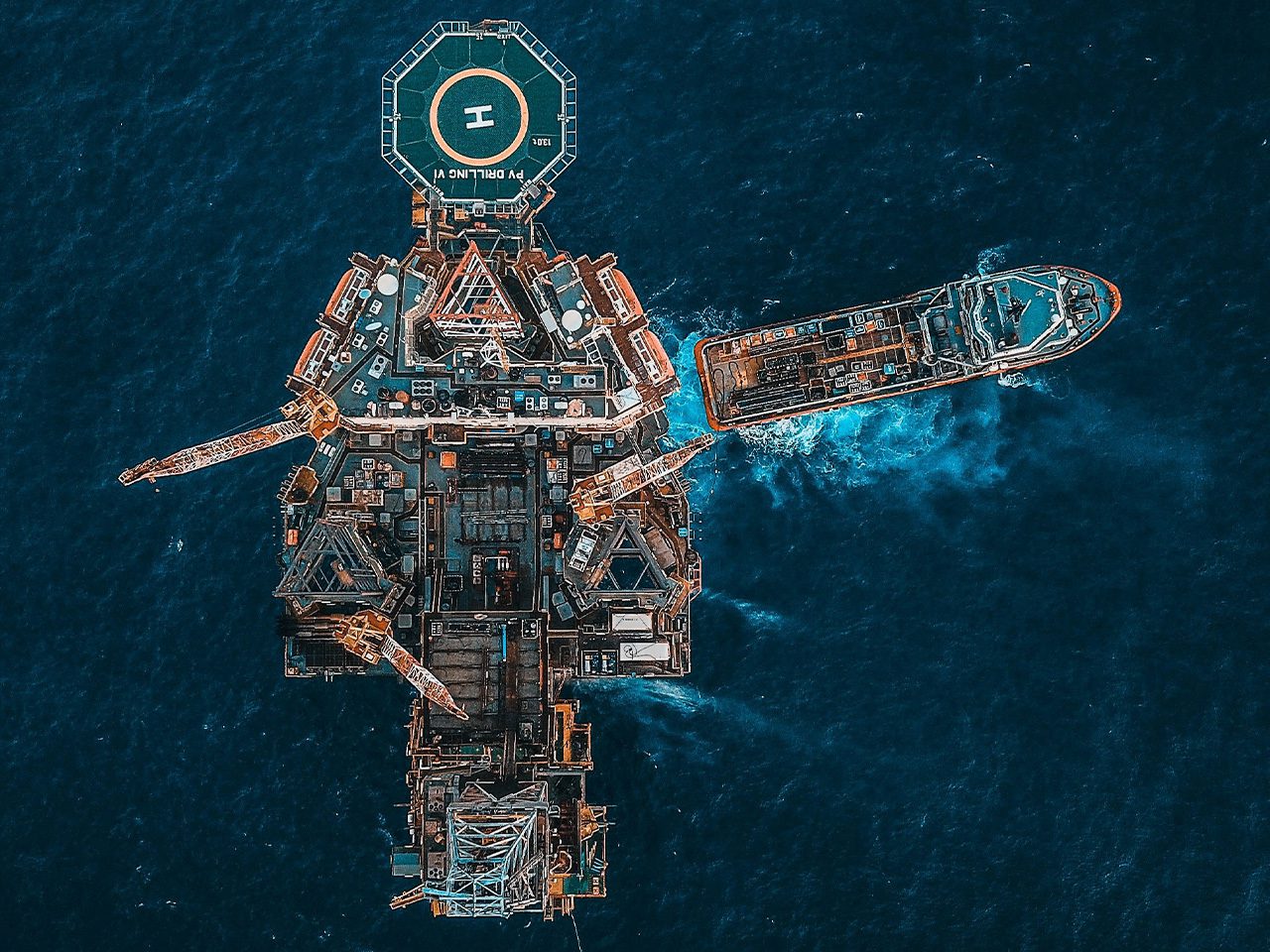 Aerial view of a large offshore platform with a helicopter landing pad and a ship nearby, floating on dark blue ocean water.