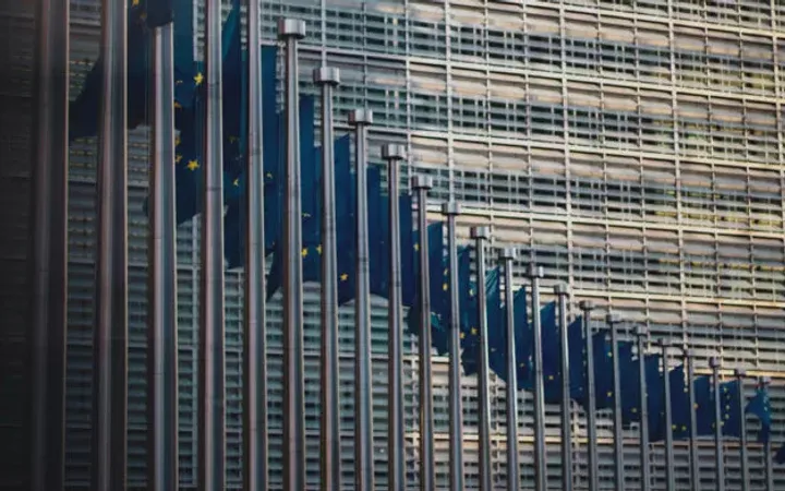 Close-up of European Union flags waving outside a modern glass building. The flags are lined up vertically, with the cityscape reflecting on the glass.