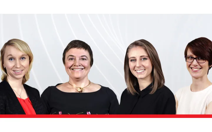 Four people standing together indoors, smiling and facing the camera, with a light background featuring white swirl patterns.