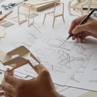 Hands sketch architectural drawings on large white sheets, surrounded by wooden chair models, blocks, and drafting tools on a workspace.