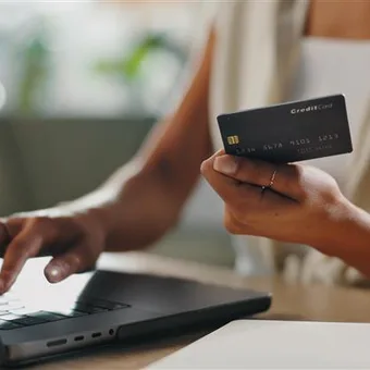 A person with long wavy hair and black-rimmed glasses, smiling at the camera, wearing a black blazer over a white top and a pendant necklace.