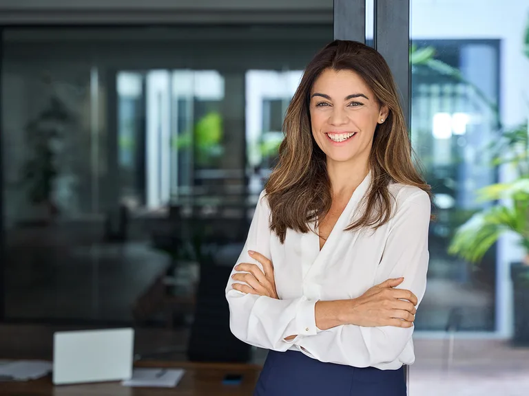 Smiling person with long brown hair, wearing a white blouse and dark skirt, arms crossed in a bright modern office with glass walls.