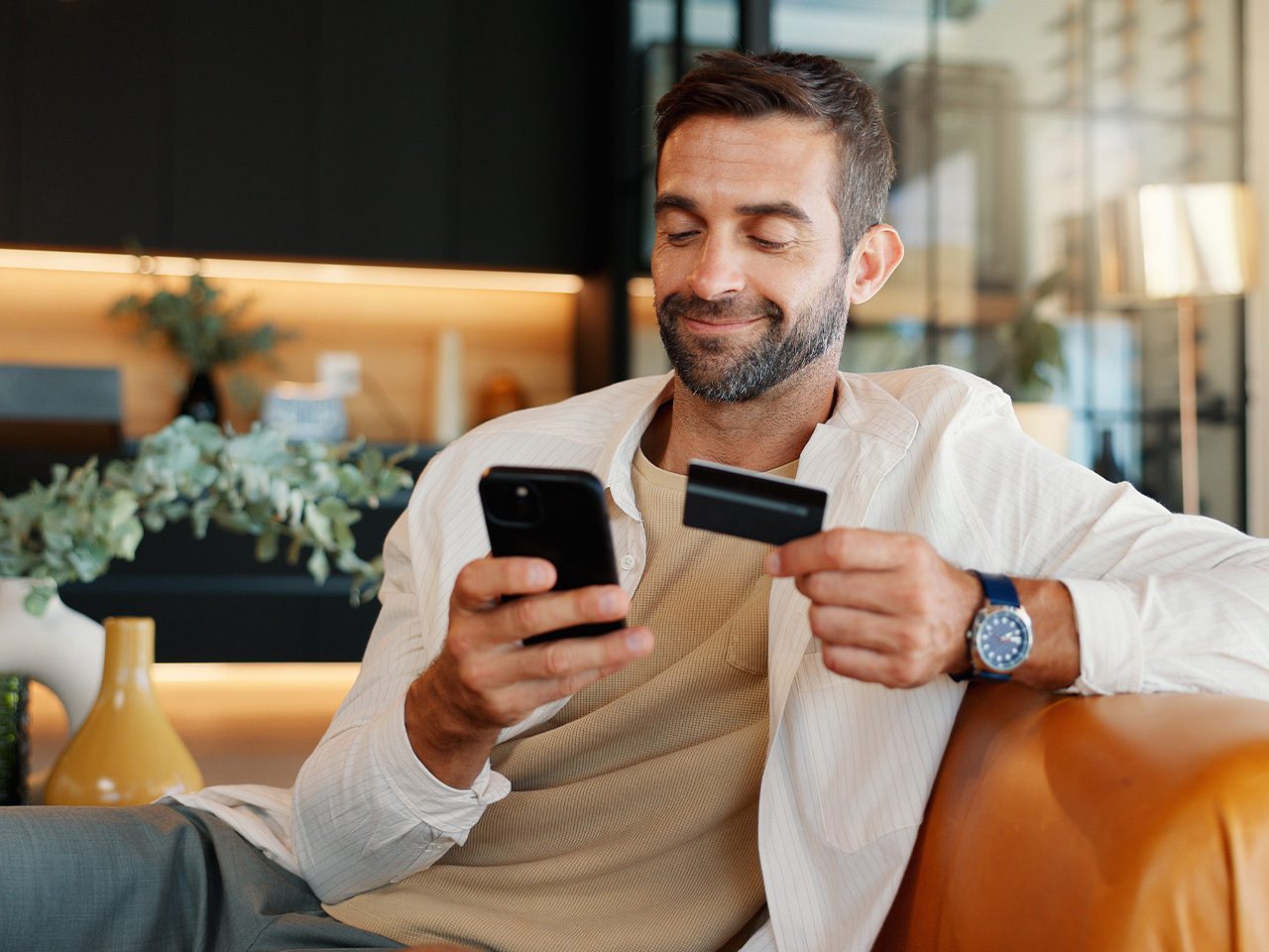 A person with short hair and a beard sits on a leather sofa, smiling, holding a smartphone and a credit card in a modern, plant-filled room.
