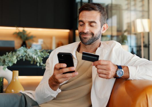 A person with short hair and a beard sits on a leather sofa, smiling, holding a smartphone and a credit card in a modern, plant-filled room.