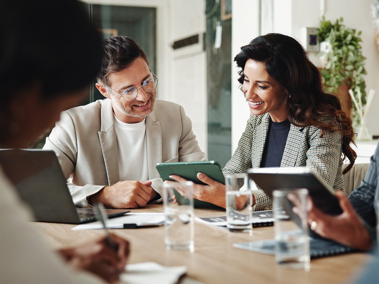 Two colleagues sit at a wooden table in a bright office, smiling as they look at a tablet together, with laptops and water glasses nearby.