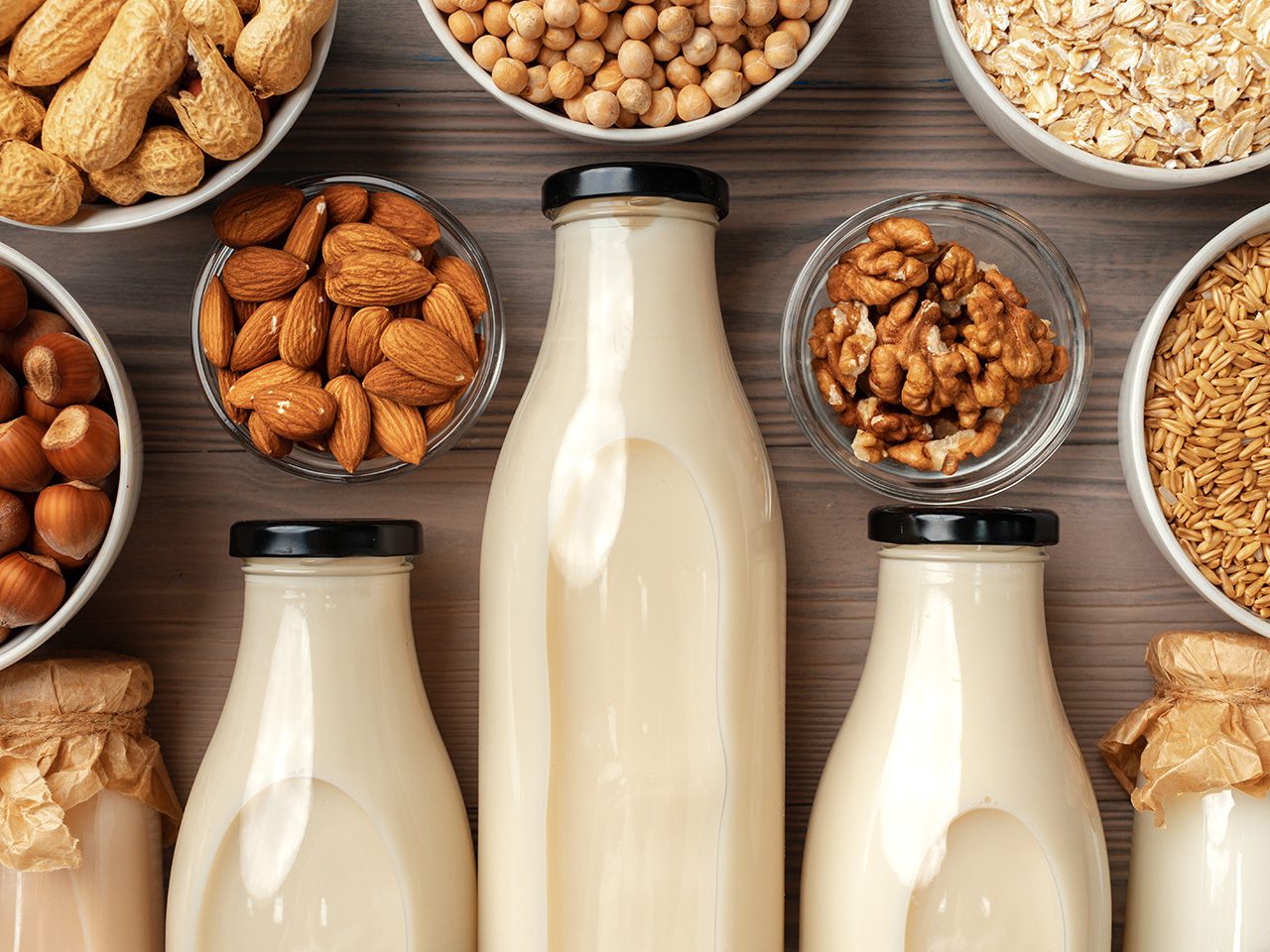Top-down view of three bottles of creamy nut milk surrounded by bowls of almonds, hazelnuts, walnuts, peanuts, oats and chickpeas.
