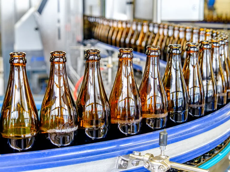 Amber glass bottles on a blue curved bottling conveyor in an industrial factory, with reflections from overhead lights on the bottles.