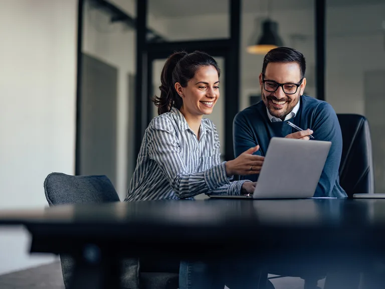 Two people sit at a conference table with a laptop, smiling as they review something together; glass walls and a pendant lamp in a modern office.