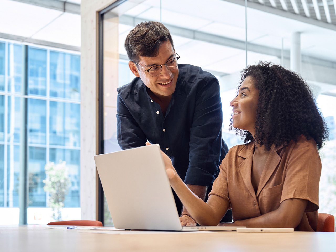 Two people collaborate at a table with a laptop in a bright, glass-walled office, one standing and smiling as they discuss the screen.