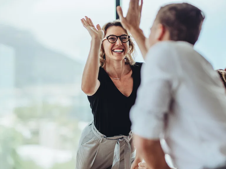 Two coworkers high-five in a bright office; the person facing the camera wears glasses, a black top, and light gray pants, smiling broadly.