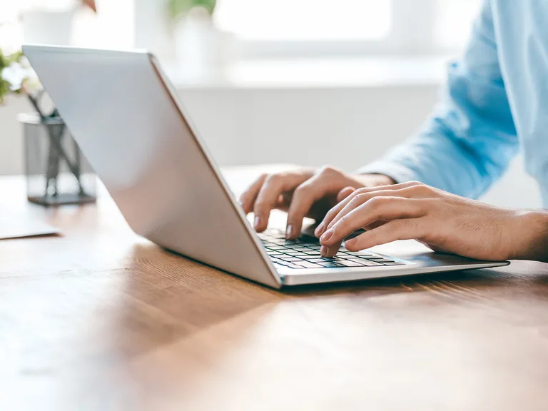 Person typing on a slim silver laptop on a wooden desk; hands on the keyboard, wearing a light blue shirt in a bright, airy room.