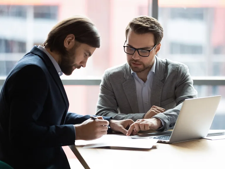 Two people in business attire sit at a desk by a large window, reviewing documents; one writes while the other points at pages, with a laptop nearby.
