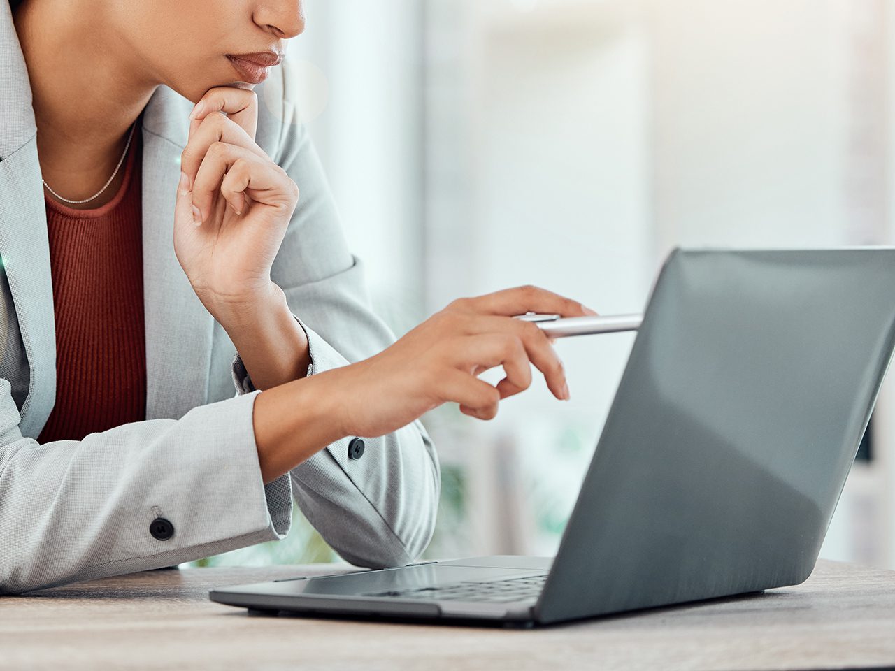 Person in a light gray blazer and red top uses a stylus on a laptop, resting their chin on a hand in a bright office setting.