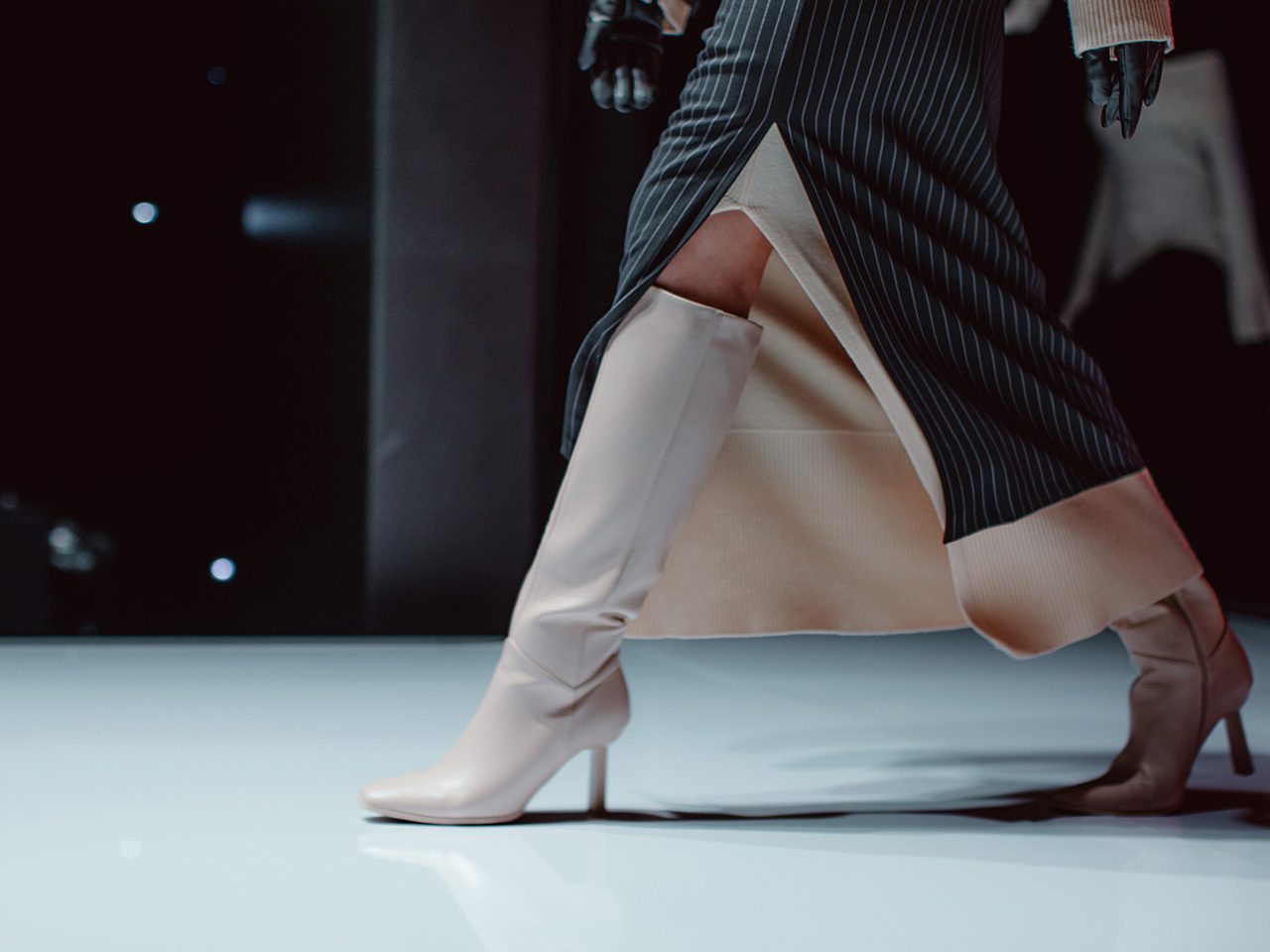 Close-up of a model walking the runway in pale knee-high boots and a pinstriped coat with a side slit on a glossy white catwalk.