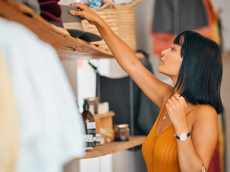 Person with shoulder-length black hair wearing an orange sleeveless top reaches up to grab a hat from a display shelf in a shop, with jars and baskets in the background.