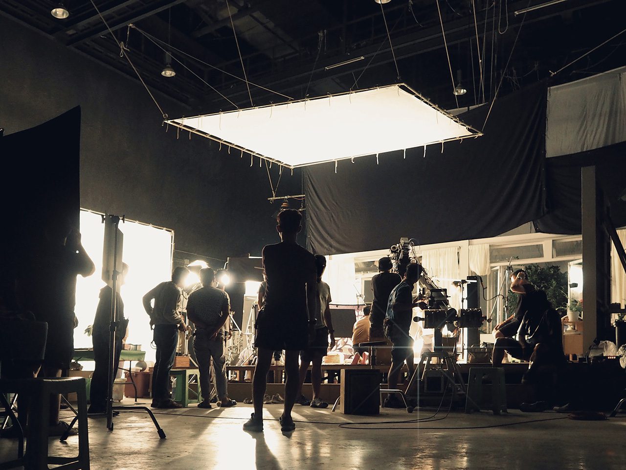Film studio set with crew and cameras, bright overhanging light, cables and equipment, silhouettes of people at work.
