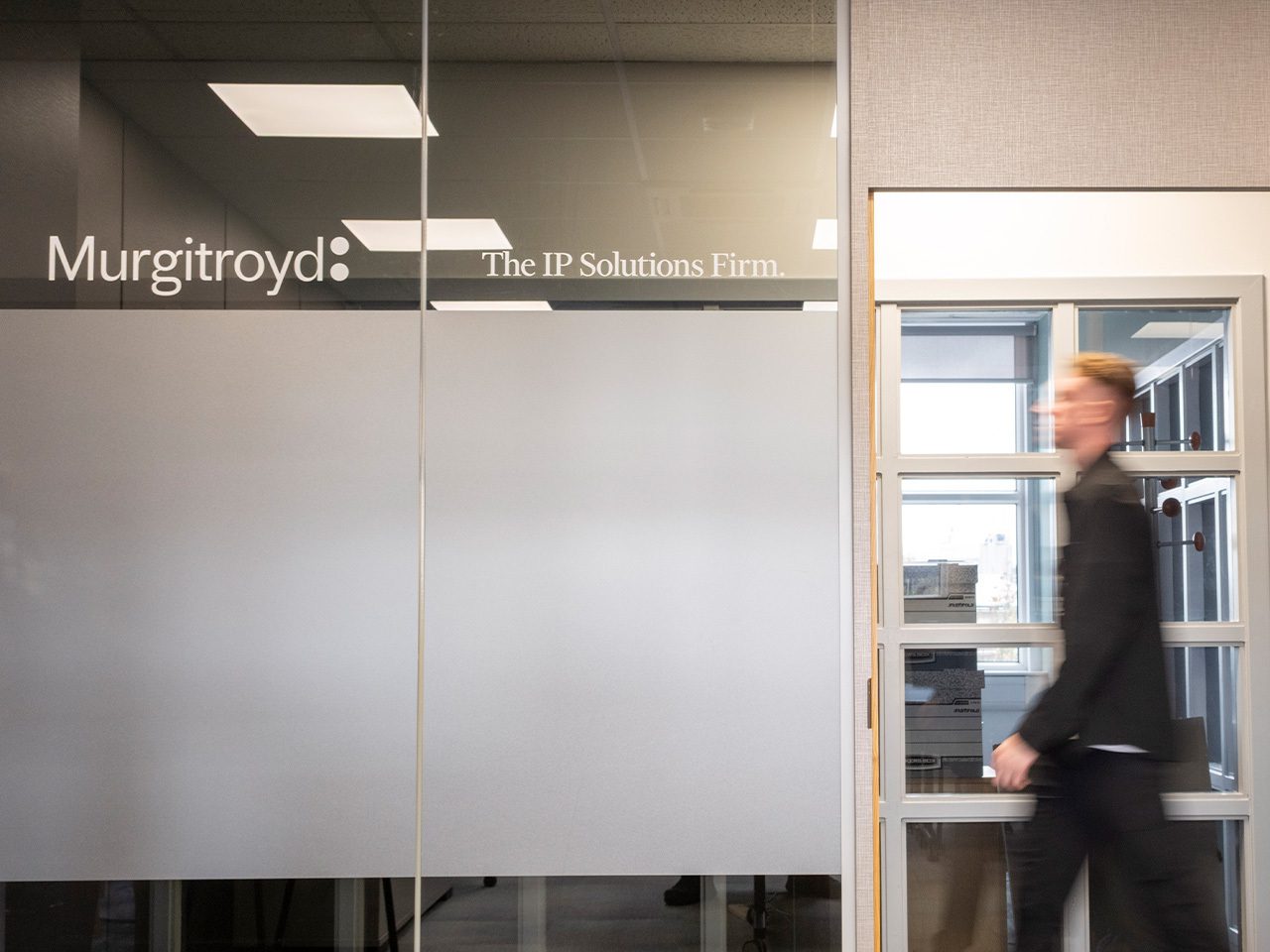 Murgitroyd glass wall with 'The IP Solutions Firm' text; a blurred person passes a glass door in a modern office interior.