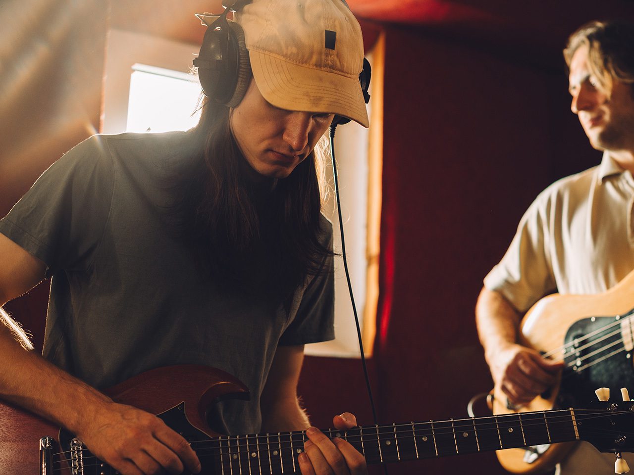 Two musicians in a warm recording studio: one with a beige cap and headphones playing an electric guitar; the other in a light polo playing bass.