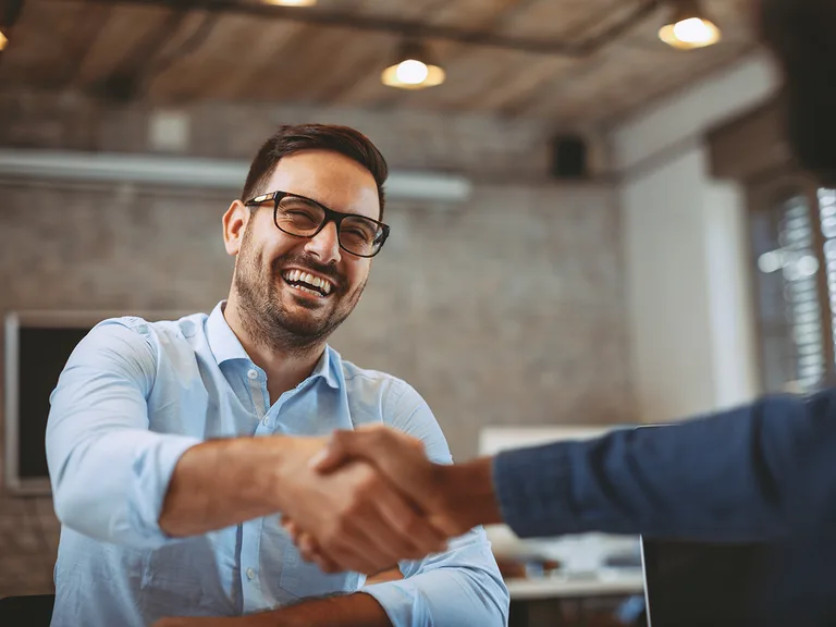 A smiling person with dark hair and glasses wearing a light blue shirt shakes hands with another person in a brick-walled office.