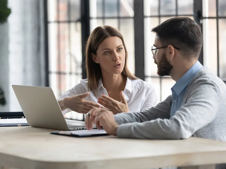 Two people sit at a bright office desk, one speaking with hand gestures while the other listens, with a laptop and papers on the table.