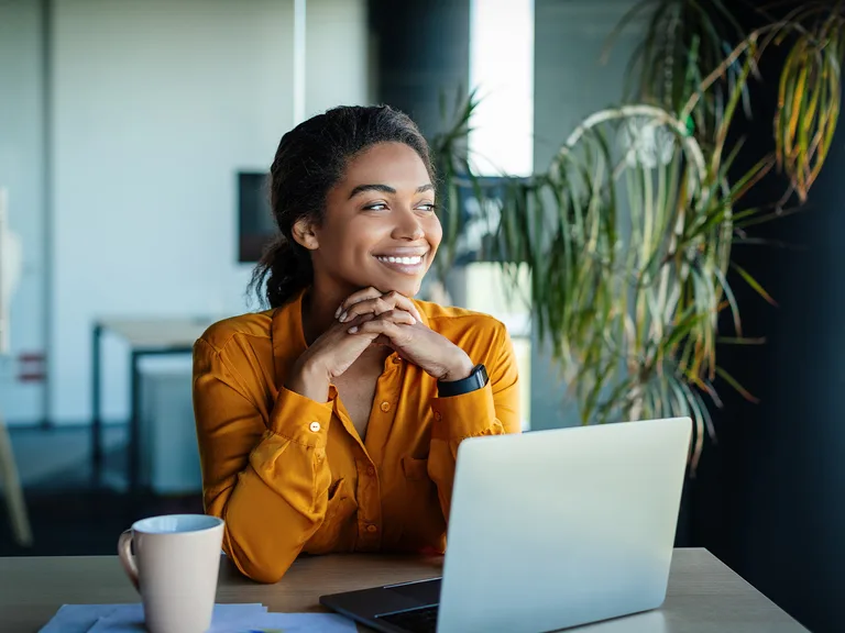 A person with dark hair wearing an orange shirt smiles with hands folded under the chin at a desk with a laptop, mug and plants in the background.
