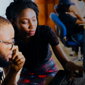 Two young adults working together on a laptop in a shared office space, with others working at desks in the background.