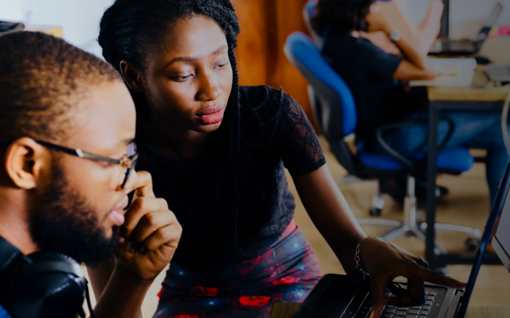 Two young adults working together on a laptop in a shared office space, with others working at desks in the background.