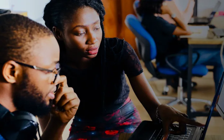 Two young adults working together on a laptop in a shared office space, with others working at desks in the background.