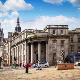 A grand historic building with columns and domed architecture, situated on a cobbled street with parked cars, pedestrians, and sculptures in front, under a partly cloudy sky.